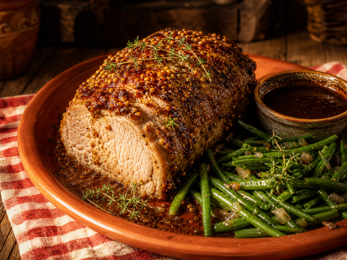 Palette de porc à la moutarde, cuite au four, servie avec des haricots verts et une sauce, sur un plat en terre cuite.